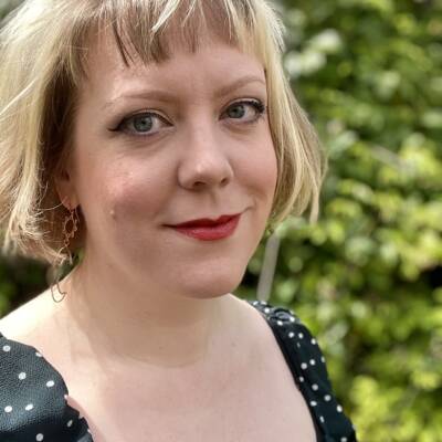 A head and shoulders image of Hazel Gardner, a white woman with a blonde bob, fringe, and dark eyes looking askance at the camera and smiling slightly. She wears red lipstick and a black and white polkadot top with puffy sleeves. Set against a green hedge.