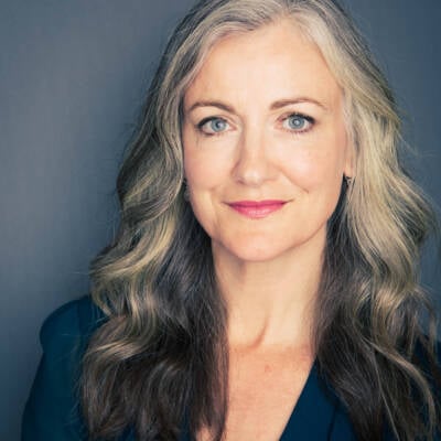 A white woman with grey hair, blue eyes, and pink lipstick, standing against a blue background and looking at the camera