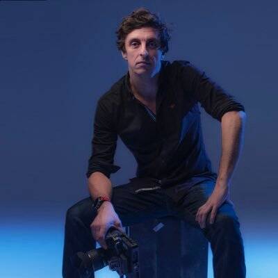 Gez Medinger, a long-limbed white man with dark curly hair, sitting on a black box against a dark blue background. He has one hand on his thigh, the other holding a professional camera, and looking straight at the camera taking his picture.