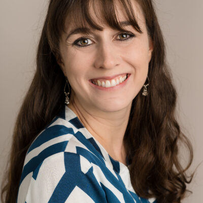 Megan Hopkins, a white woman with long brown hair and a fringe is smiling at the camera over he shoulder, in a white and dark blue geometric top