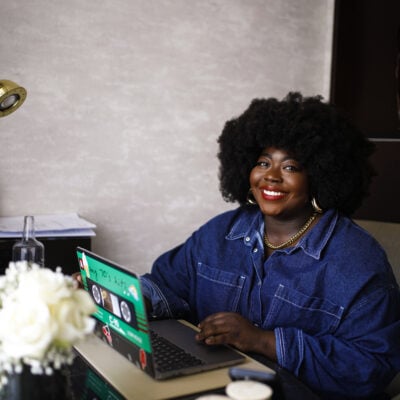 Stephanie Yeboah, a Black woman with red lipstick and an afro, is sitting at a desk with her laptop open, and smiling at the camera. She is wearing a dark-wash jean jacket and the wall behind her is grey. There is a small vase of white roses on the table next to her.