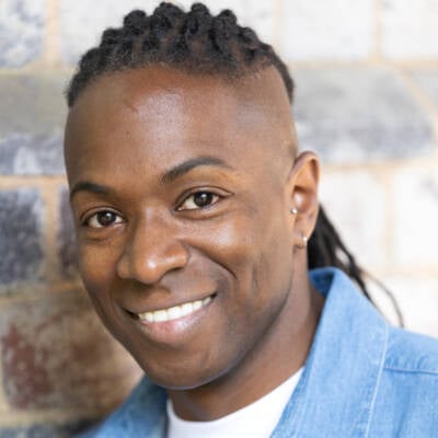 Nigel Clarke, a Black man with close braids, is wearing a white t-shirt and blue collared shirt, open, and smiling at the camera from an oblique angle.