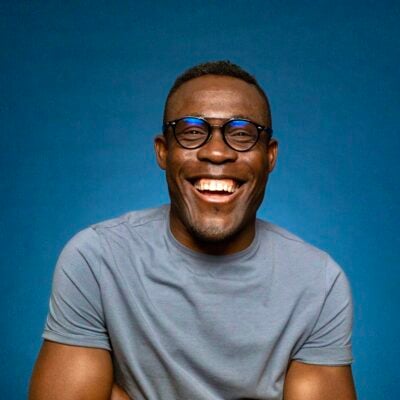 Headshot of Peter Komolafe, a black man with glasses in a grey t-shirt smiling widely at the camera. He is set against a blue background.